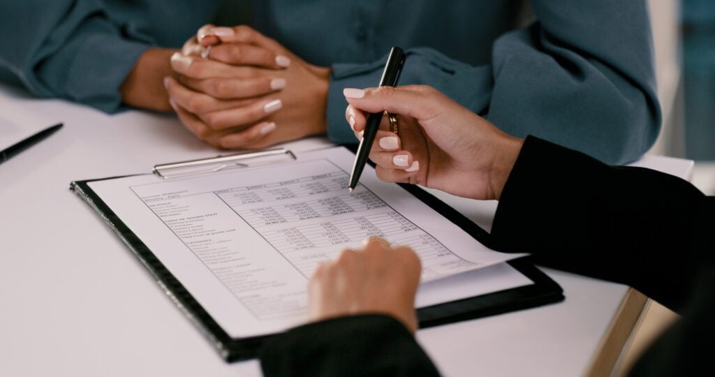 Two people sit at a table reviewing a document with a table of numbers on a clipboard; one person holds a pen and gestures toward the document while the other has hands folded.